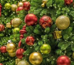 A beautifully decorated Christmas tree filled with festive ornaments. The tree is adorned with shiny red, gold, and green baubles, along with small golden gift boxes and elegant swirled designs on some ornaments.